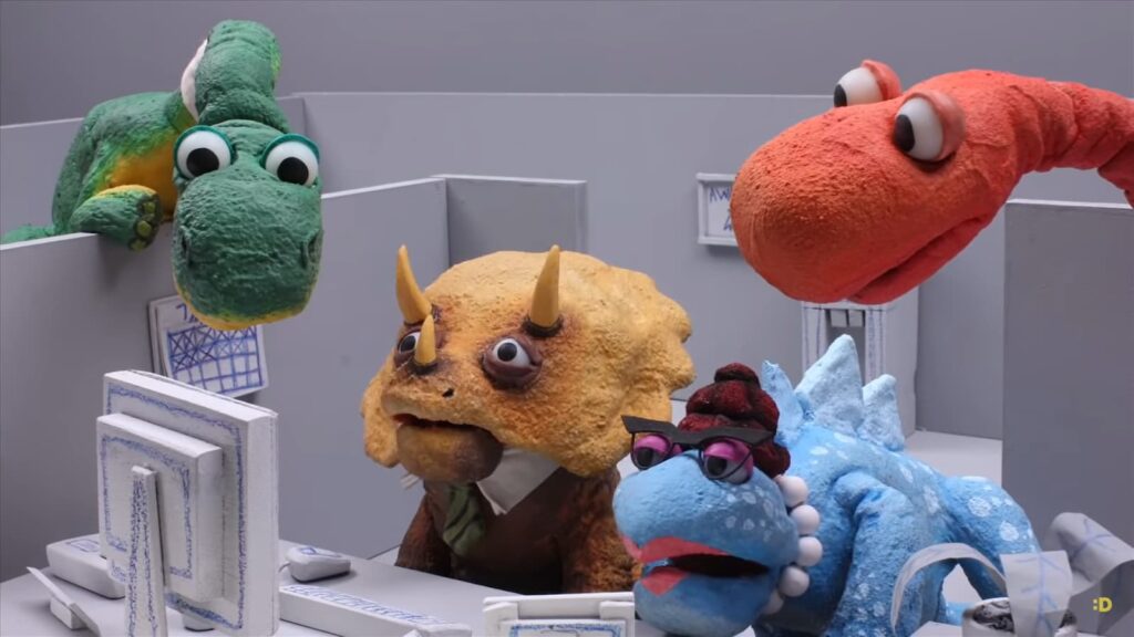 Colorful dinosaur puppets in an office setting with computers, one wearing glasses and a hat